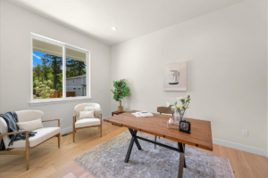 Bright home office with a wooden desk, two cream chairs, a gray rug, and a window showing trees outside.