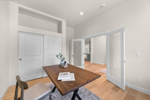 Bright home office/dining area with a wooden table, a chair, and white double doors; glass-panel doorway opens to the kitchen hallway.