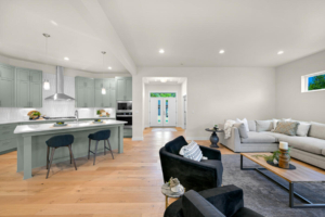 Open-concept living area with a pale green kitchen, island, and bar stools; bright, airy seating area with gray sofa and black chairs outside the kitchen.