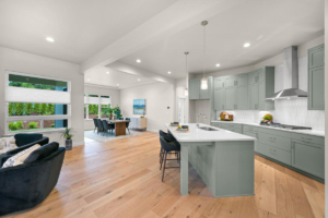 Open-concept kitchen with sage-green cabinets, white island, and stainless range hood; dining area visible beyond.
