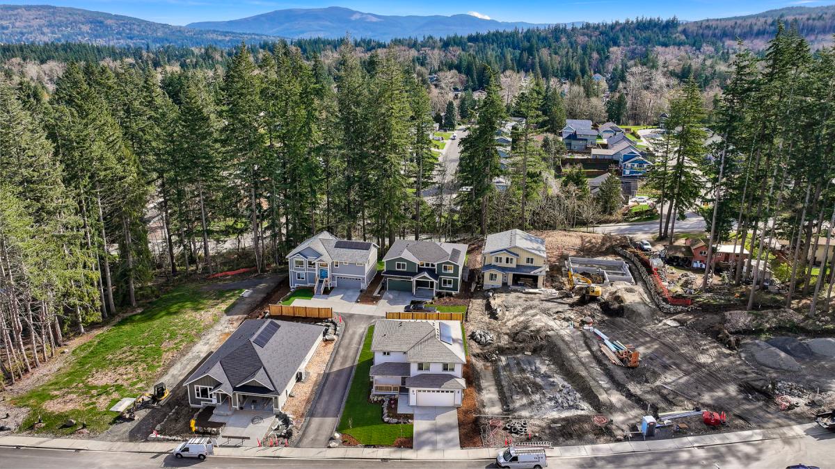 Aerial view of a suburban street with several houses under construction beside a dense pine forest and distant mountains.