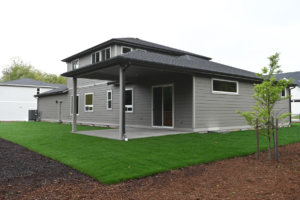 Modern gray two-story house with a covered patio, sliding glass door, and a well-kept green lawn in the backyard.