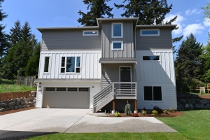 Modern gray two-story home with a front porch, stairs, and a two-car garage, surrounded by trees and a lawn.