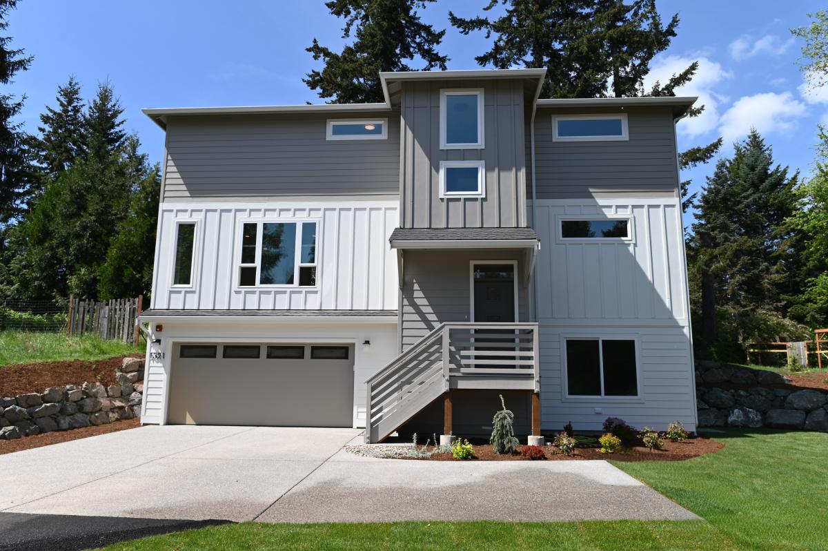 Modern gray two-story home with a front porch, stairs, and a two-car garage, surrounded by trees and a lawn.