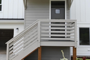 Front entrance of a modern townhome with a raised wooden porch, stairs, and gray siding, surrounded by mulch and small shrubs.