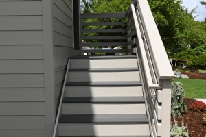 Exterior staircase with gray steps and white railing leading to the front door of a gray house; shadow falls on the left side, with landscaped yard nearby.
