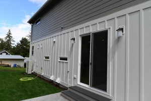 Backyard view of a modern gray-siding house featuring a sliding glass door and outdoor wall lights on the right side, with a green lawn and hose in the foreground.
