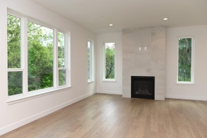 Bright living room with light hardwood floors and a modern vertical fireplace flanked by large windows showing greenery outside.