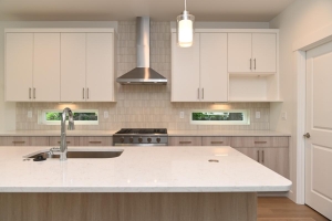 Modern kitchen with white upper cabinets, light wood lower cabinets, a marble island, and a stainless steel sink with faucet plus a gas range under a stainless hood.