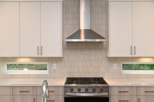 Modern kitchen with a stainless steel range and vent hood, white upper cabinets, and a light tiled backsplash between two small horizontal windows.