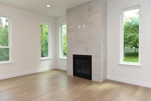 Bright living room with a tall tiled fireplace chimney in the center, flanked by three large windows and light hardwood flooring.