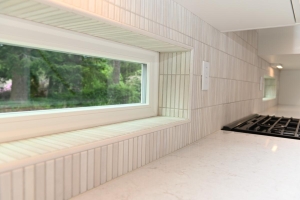 A modern kitchen with a wide horizontal window set in a tiled wall above a white marble countertop and a black gas stove on the right, overlooking greenery outside.