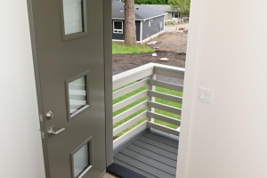 Interior view of a gray front door opening to a small balcony with white railing and gray decking, neighborhood houses visible outside.
