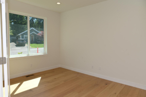 Empty room with light wood floor and a large window letting in sunlight; view of a suburban house outside.
