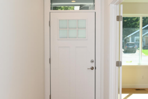 White front door with a six-pane window, set in a bright hallway with light hardwood floors and an adjacent sunlit room to the right (transom window above).
