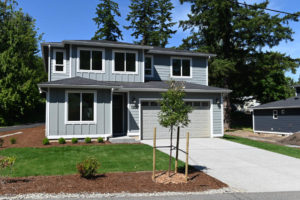 Two-story gray suburban house with an attached two-car garage, front yard, and a young tree in mulch.