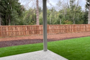 Covered backyard patio with a concrete slab, grey support post, and a wooden fence beyond a strip of green lawn. A tree line and mulch path are visible in the background.