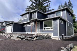 Two-story blue house under construction with large front window, gravel driveway, and rock retaining wall.