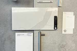 Flat lay of assorted tile samples and a metal door handle arranged on a floor, with a small vase of white flowers in the top-left corner.