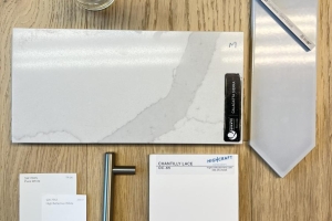 Flat lay of interior decor samples on a wooden table: marble tile, metal handle, swatches, envelope, and a small vase with white flowers.